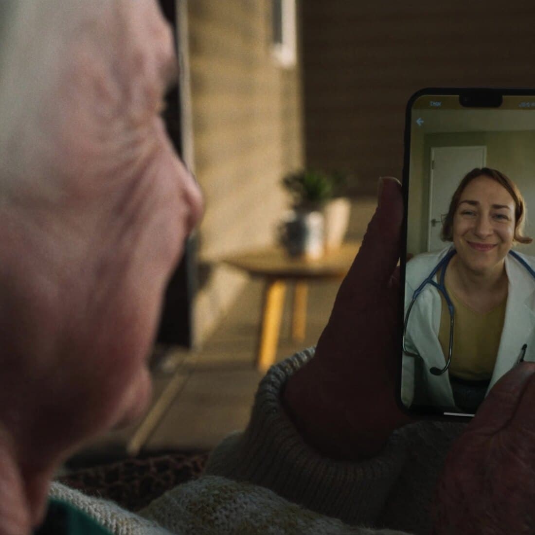 An older man at home on a video call with his clinician, who smiles from the phone screen